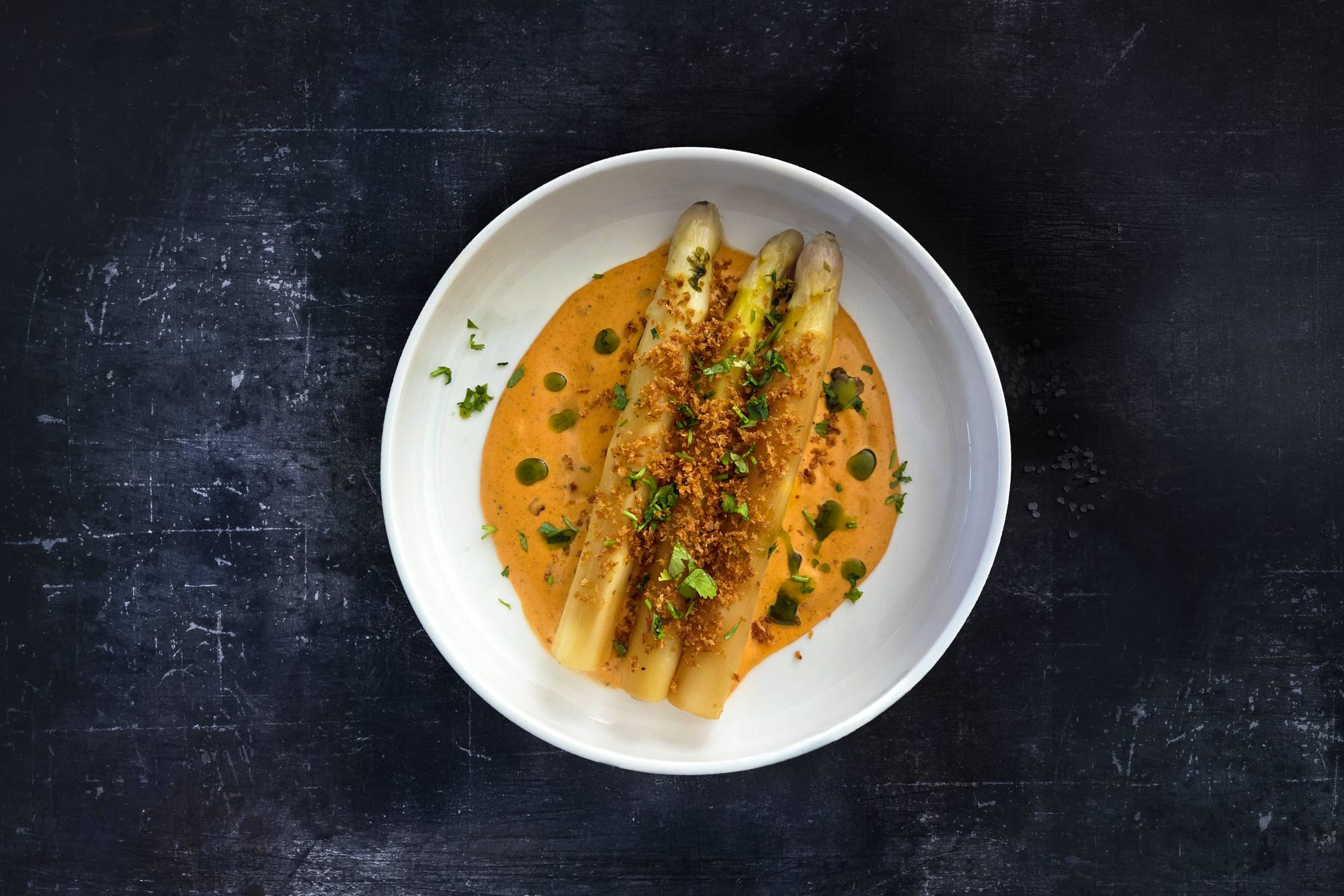 Sous-vide asparagus with miso butter & gochujang hollandaise White sous-vide asparagus in a creamy gochujang hollandaise, garnished with herbs, miso butter and a crispy topping, served in a white bowl.
