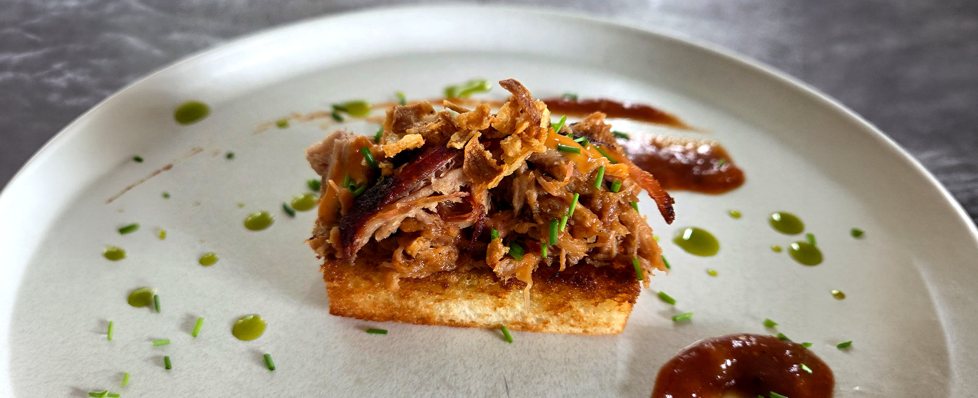 Pulled Pork auf knusprigem Brot mit Röstzwiebeln, Schnittlauch, Sauce und Kräuteröl auf einem weißen Teller.