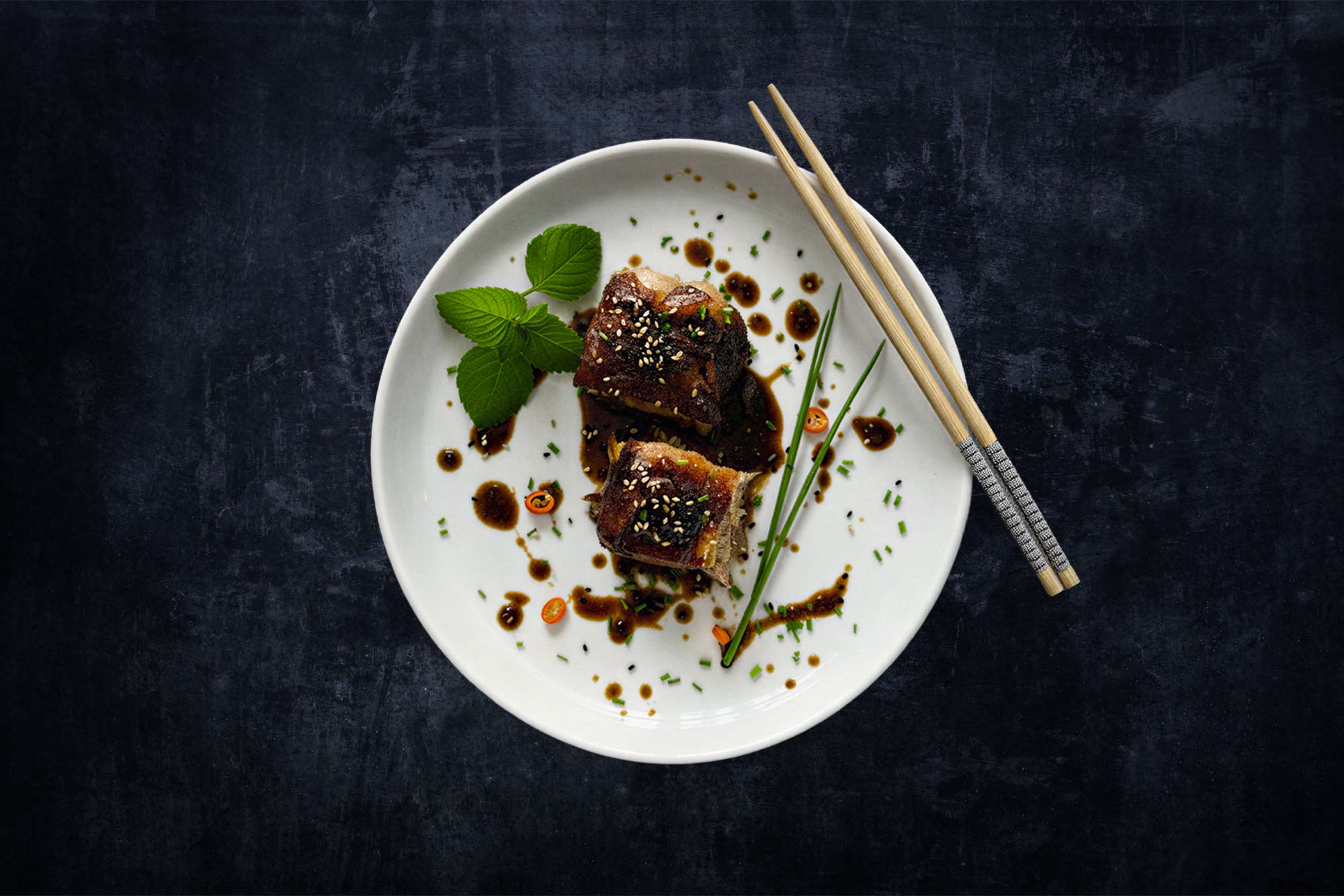 Two pieces of meat with a dark glaze are arranged on a white plate with herbs, sesame seeds, chilli and chives. To the right of them, chopsticks lie on a dark surface.