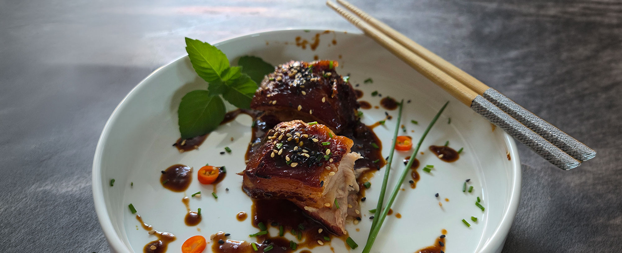 Two pieces of glazed pork belly are arranged on a white plate with a dark sauce, sesame seeds, chilli, chives and fresh herbs. Chopsticks lie at the edge of the plate.
